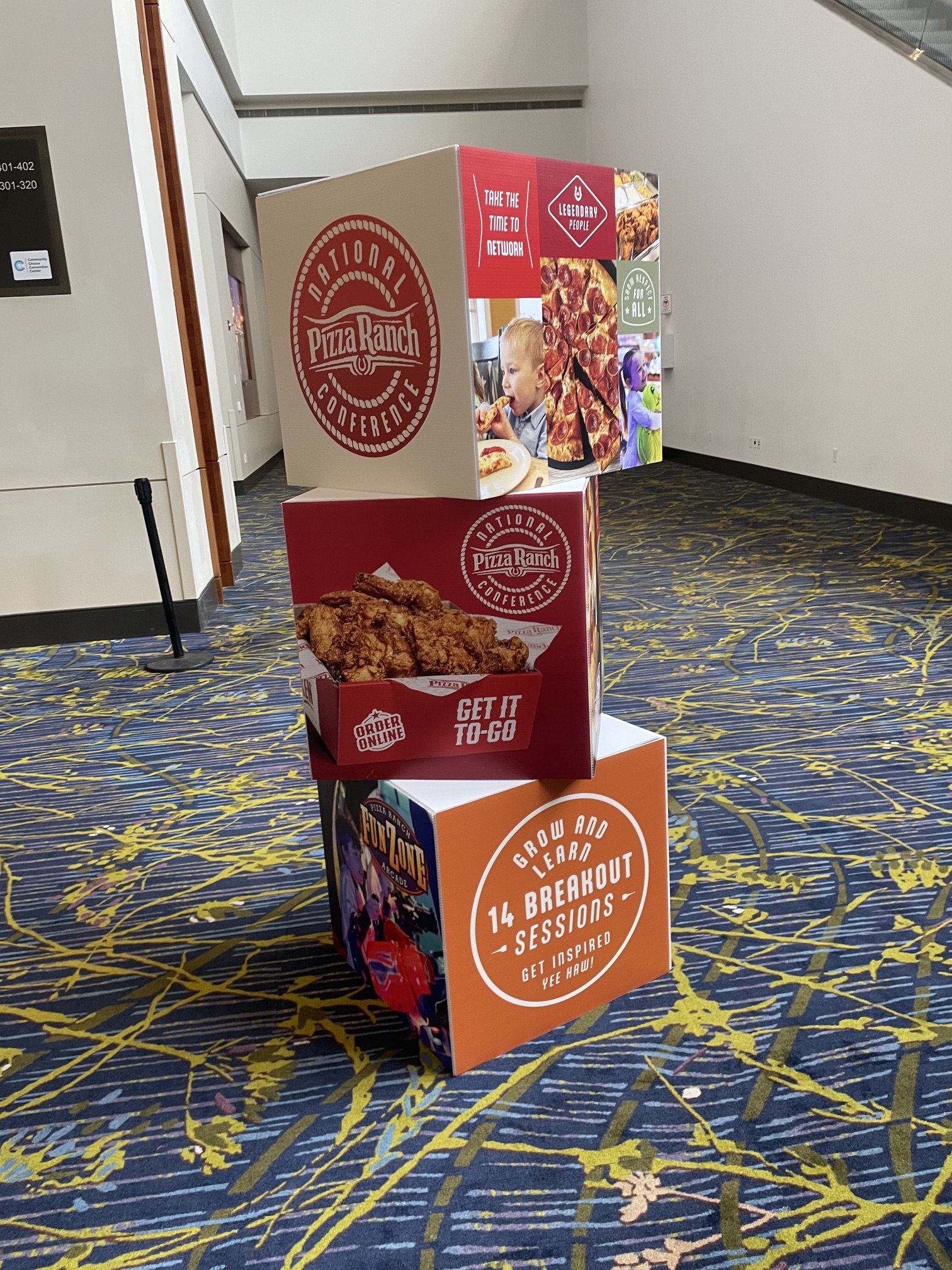 Display at a conference promoting Pizza Ranch, featuring images of pizza and fried chicken, with slogans about networking, breakout sessions, and online ordering, on a patterned carpeted floor.