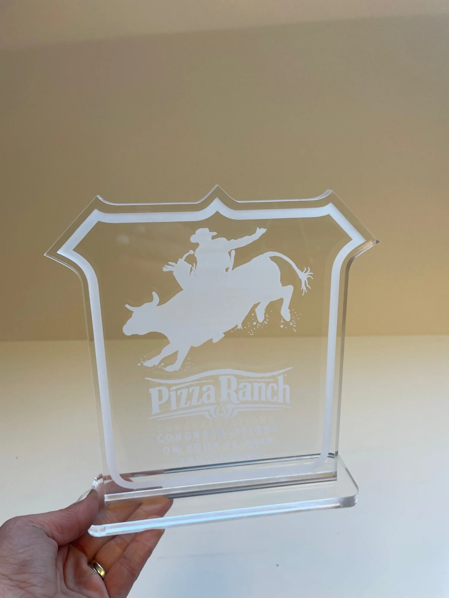 Clear acrylic sign with white graphic and text, featuring a cowboy riding a bucking bull, and the words 'Pizza Ranch' and additional smaller text, on a beige wall background.