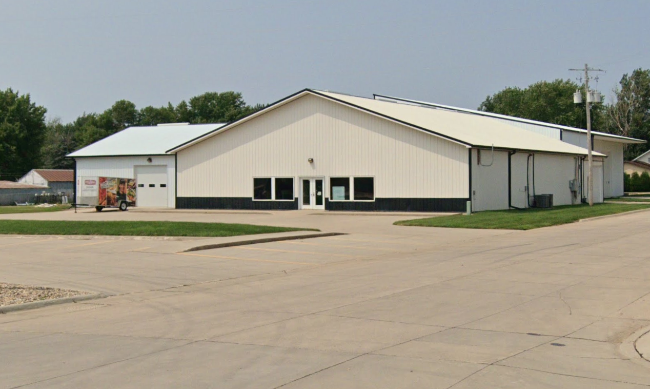 A large, white, metal building with black accents and a gabled roof, situated in an empty parking lot with some green grass nearby.
