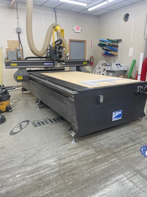 A large CNC router machine in a workshop with hoses attached, sitting on a gray carpeted floor. There are some fabric or paper sheets on the machine's work surface, and the room has shelves with rolls of material and an emergency exit sign on the wall.