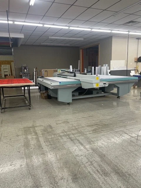 Large industrial printing or cutting machine in a workshop or printing facility with some rolls of material nearby.