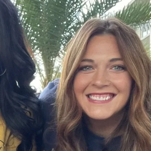 Close-up of a smiling woman with light brown hair in an outdoor setting with palm leaves in the background.