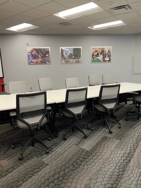 Empty conference room with six office chairs around a long white table, white walls with three framed photographs, and a gray patterned carpet.