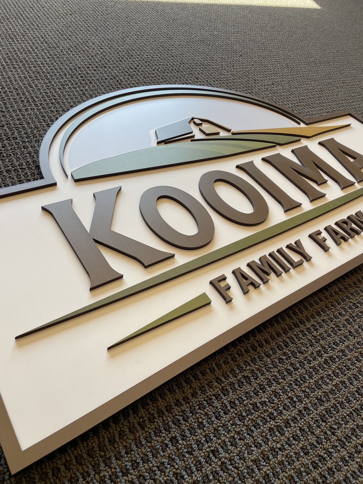 Close-up of a 3D sign or logo on a wall, with the visible text "VONT" and part of the words "FAMILY FARM" below. The sign has a green, black, and white color scheme, and is placed on a dark, textured carpeted floor.
