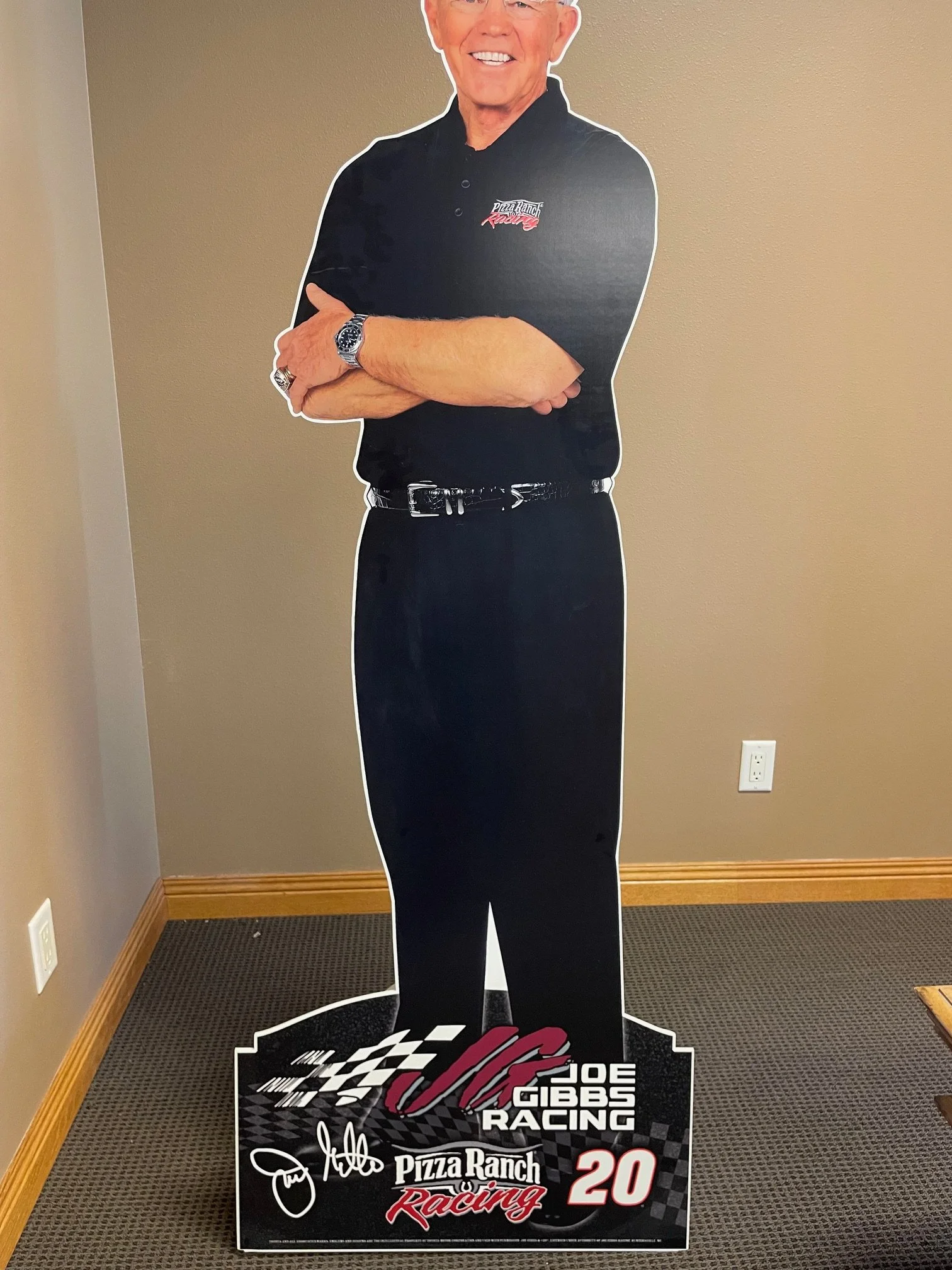 Standee of Joe Gibbs Racing team owner, featuring a smiling older man with crossed arms, wearing a black shirt with the team logo, and a bottom graphic with signatures, logos, and text including '2020'.