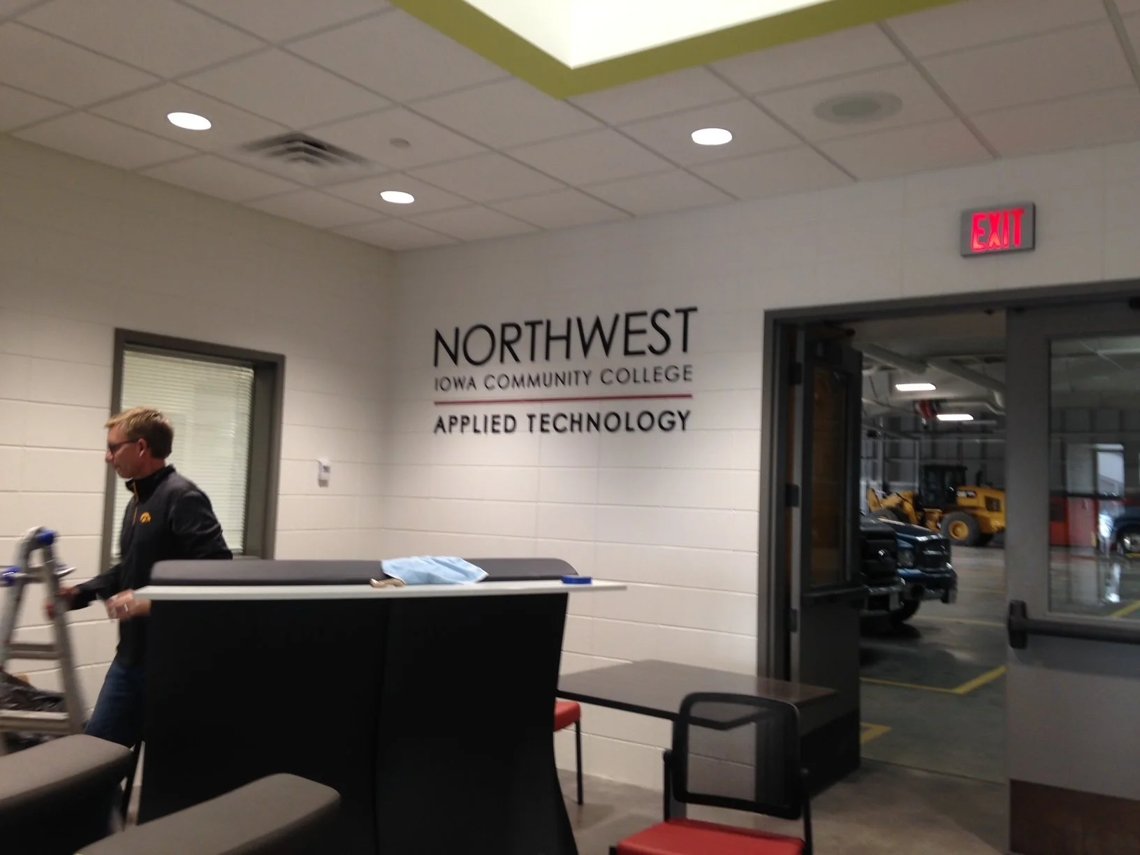 Interior of a classroom or office space at Northwest Iowa Community College Applied Technology with a visible exit door and vehicles outside, and a person in a black jacket walking by.