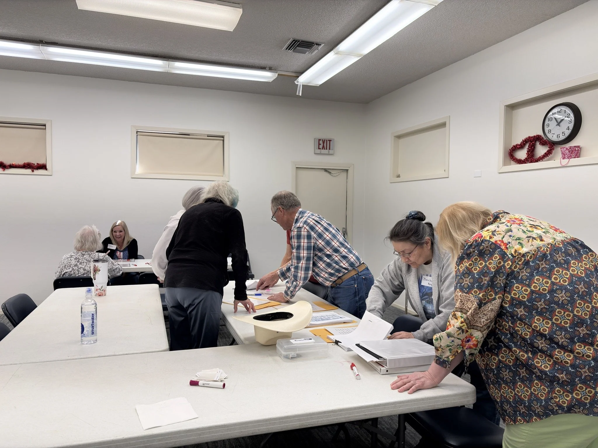 A group of people in a meeting room looking at papers on a table. There is a woman sitting at the head of the table, smiling. The room has white walls with windows and holiday decorations. There are markers and water bottles on the table.