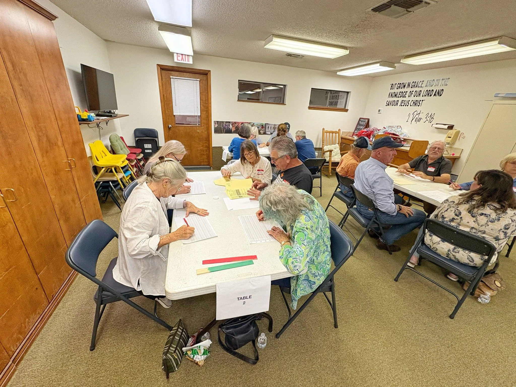 Group of elderly people sitting at tables in a meeting room, engaging with papers and discussion. The room has beige walls, windows, and a wall quote about growing in grace from 2 Peter 3:18.
