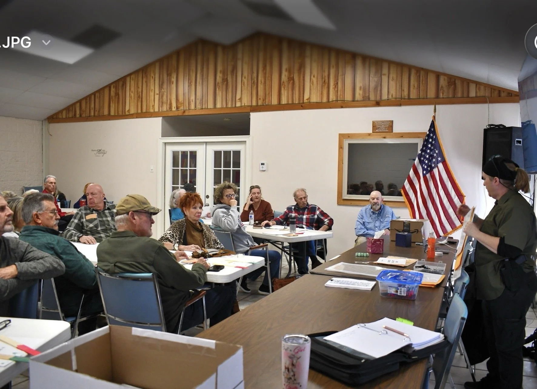 A group of people attending a meeting in a community room, sitting at tables and listening to a woman speaking at a podium. The room has a wooden ceiling, white walls, and an American flag displayed beside the speaker.