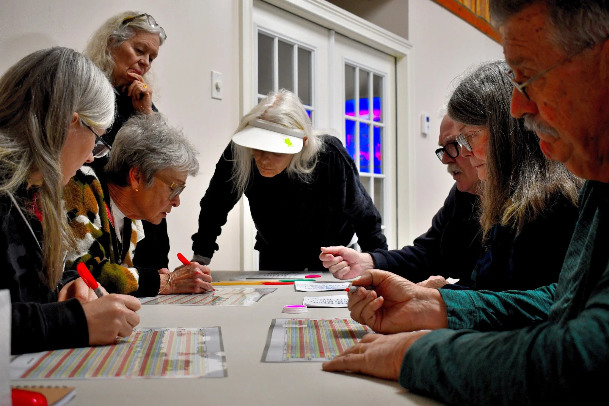 Group of women and men gathered around a table, engaged in a group activity or discussion, with printed materials and cards on the table.