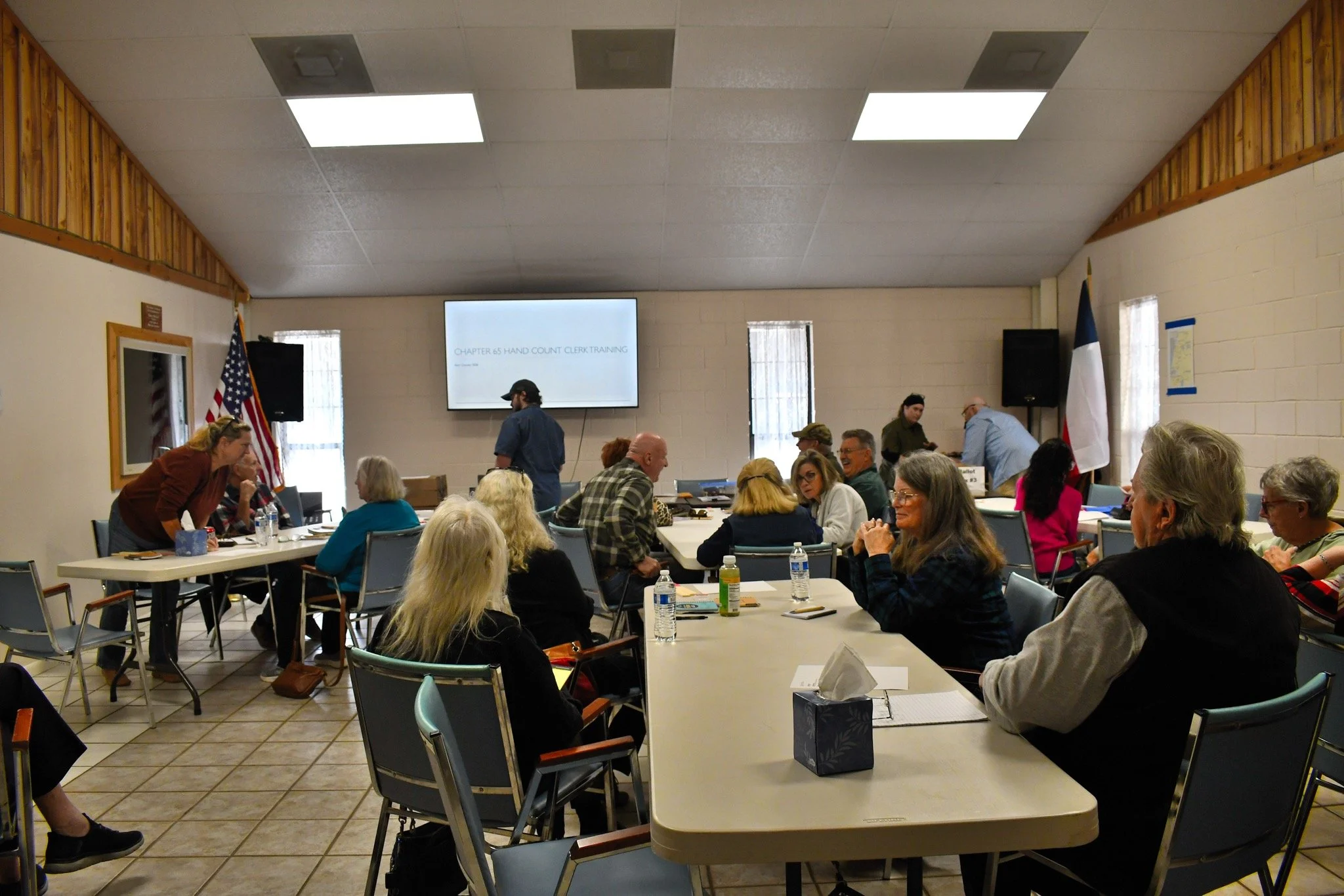 A group of people gathered in a large room with tables, participating in a training event about hand counting. There is a presentation screen on the wall displaying 'Chapter 65 Hand Count Clerk Training'. Various individuals are engaged in discussions and activities, with water bottles and other materials on the tables.