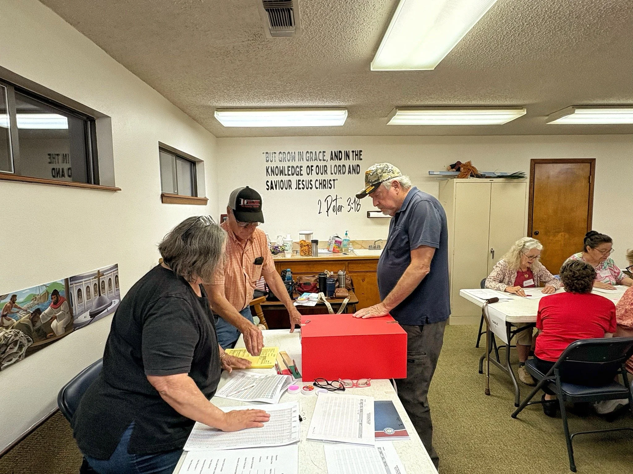 People gathered around tables with papers and documents inside a room with a religious quote on the wall.