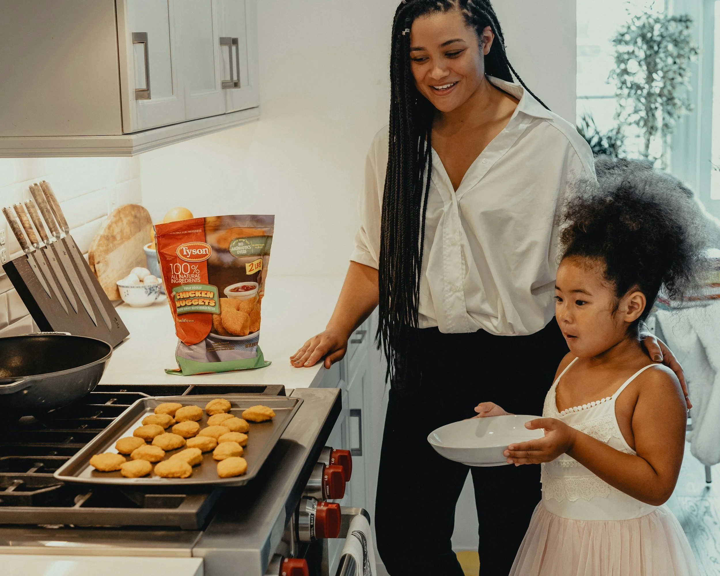 A woman and a young girl preparing food in a kitchen. The woman is smiling and wearing a white shirt, while the girl in a light dress is holding a white bowl. There is a tray of baked chicken nuggets on the stove, a bag of Tyson chicken nuggets on the countertop, and kitchen utensils nearby.
