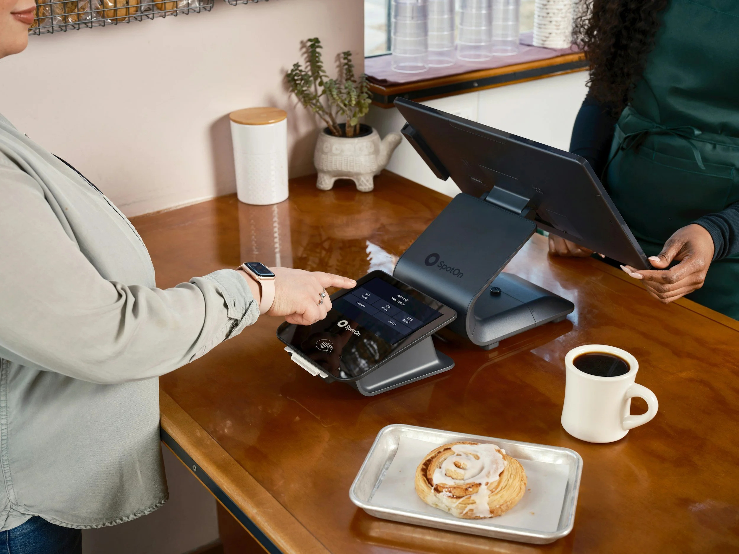 A person making a purchase at a cafe counter using a card, while a cashier processes the payment with a point-of-sale device. There are pastries, a coffee mug, and plastic cups in the background.