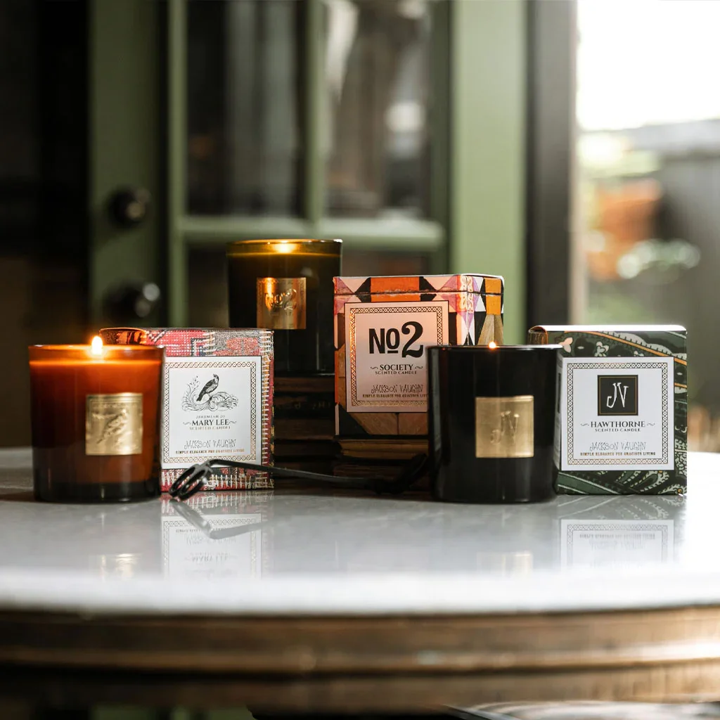 A collection of lit scented candles in decorative boxes on a marble countertop with a blurred green cabinet in the background.