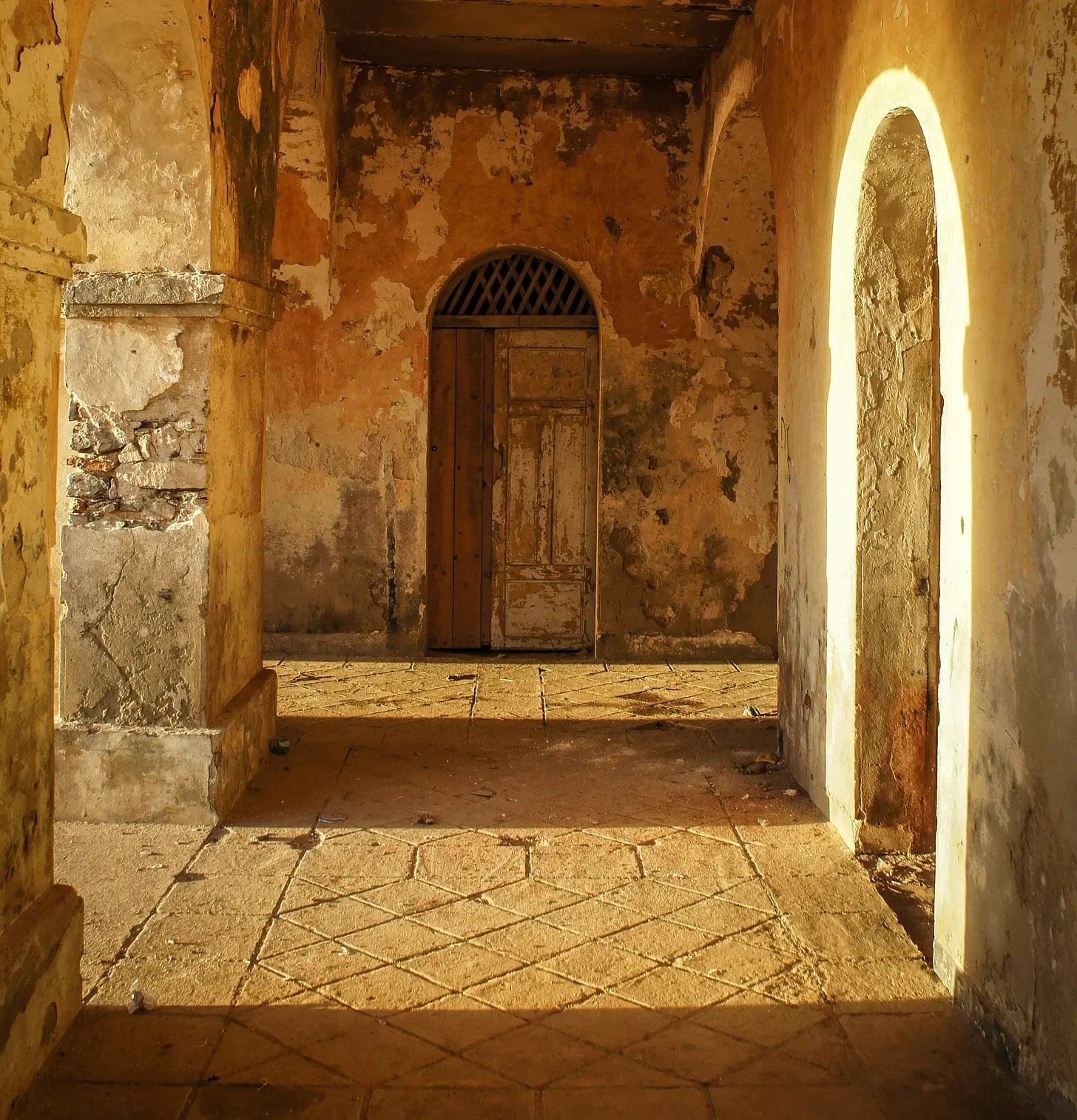 Old, weathered interior room with peeling painted walls, in warm sunlight, featuring a closed wooden door with an arched top and a small lattice window above it, and two arched doorways leading into other areas.