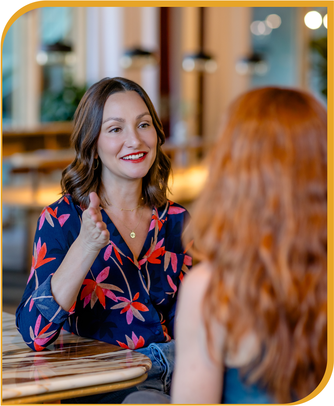 Two women having a conversation in a warmly lit casual setting, with one woman smiling and gesturing while talking.