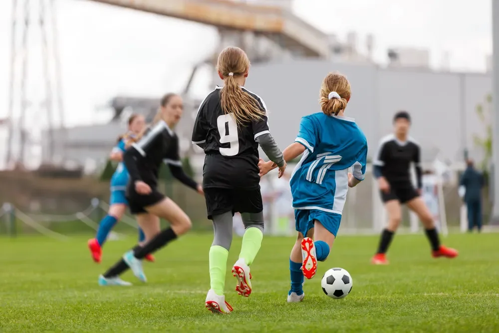 A youth soccer game with children playing on a green field, two girls chasing the ball while others watch and run in the background.