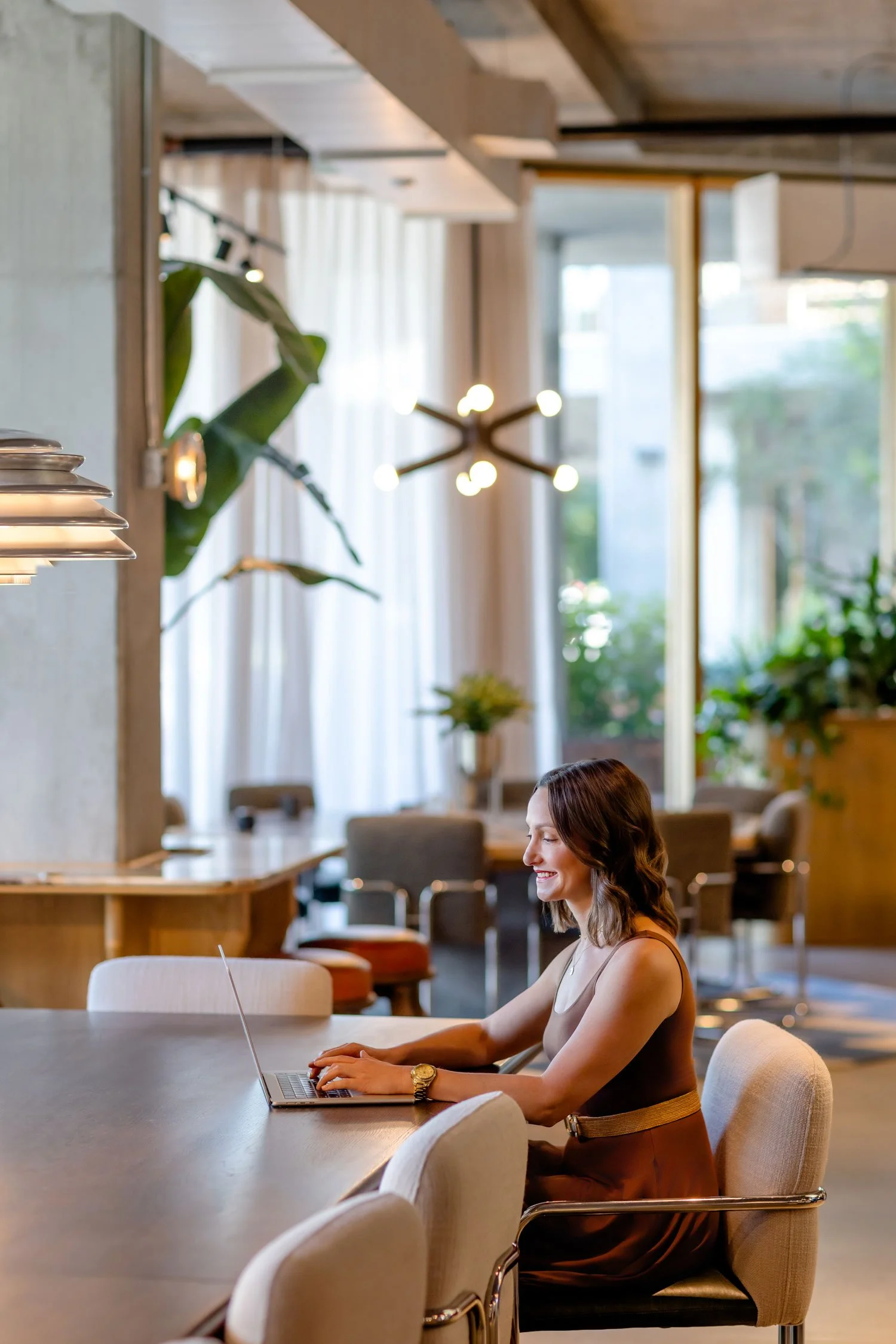 A woman with long brown hair, wearing a sleeveless top and a gold watch, sitting at a large conference table in a bright, modern, and stylish cafe or coworking space, working on a laptop with a smile.