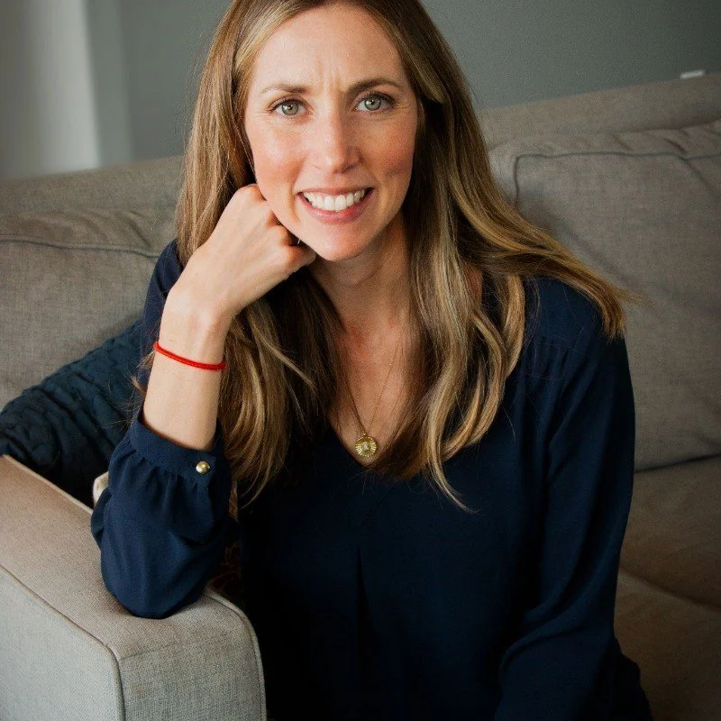 A woman with long light brown hair and green eyes smiling while sitting on a grey couch in a living room.