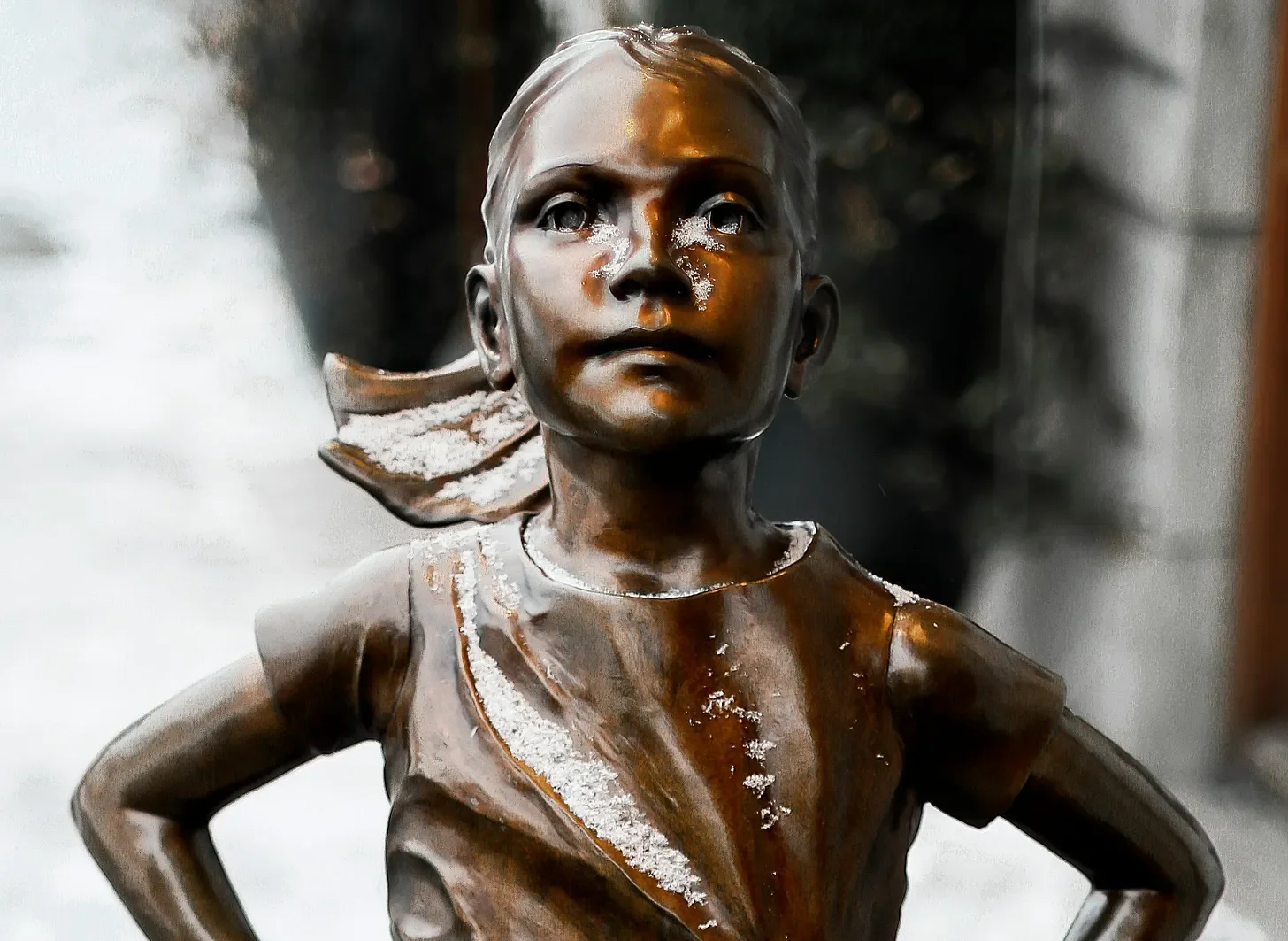 Bronze statue of a young girl with her hands on her hips, wearing a bow in her hair, with snow on her face and body.