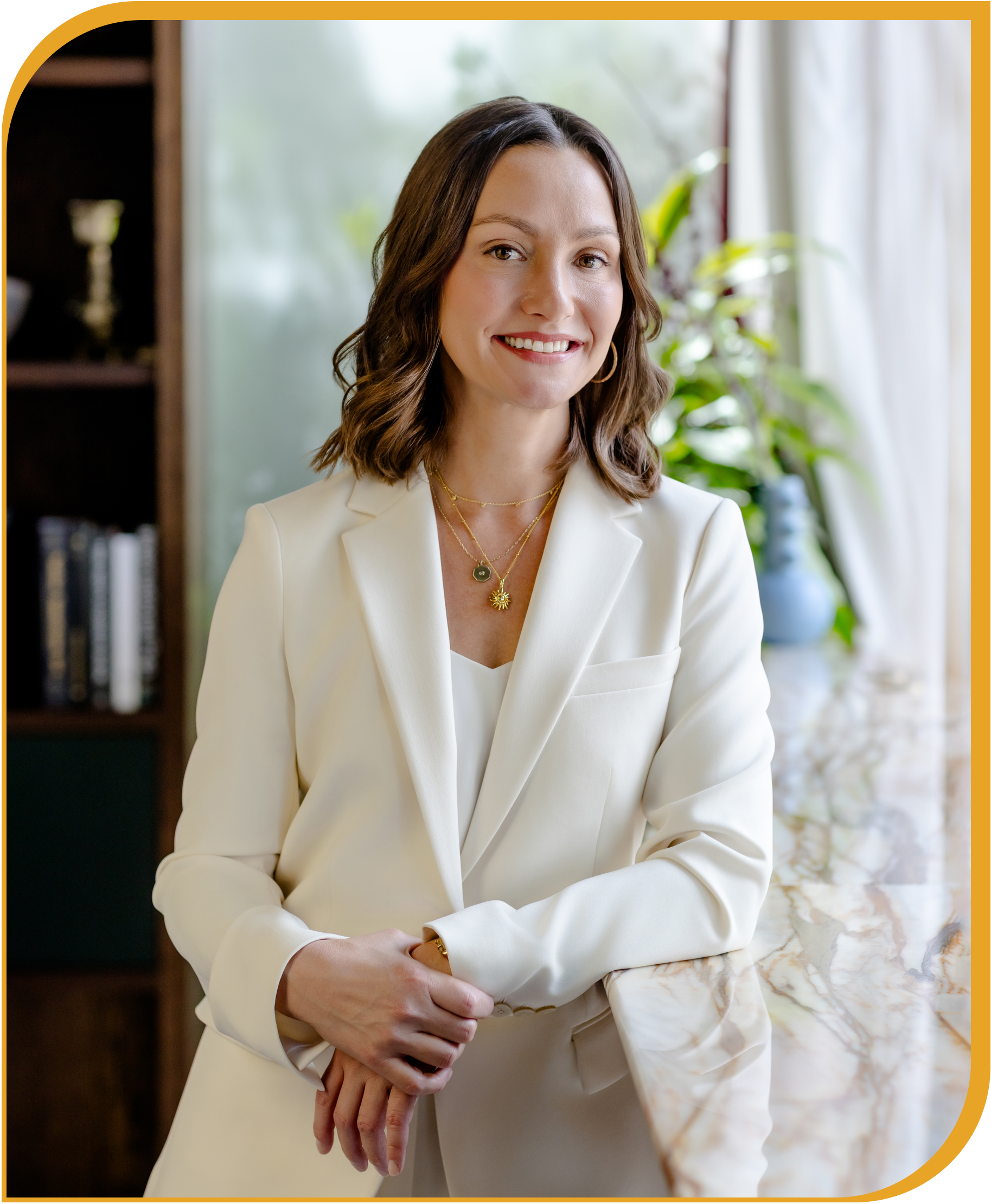 A woman with shoulder-length brown hair, wearing a white blazer and layered necklaces, sitting at a marble table in a bright room with plants and curtains in the background.
