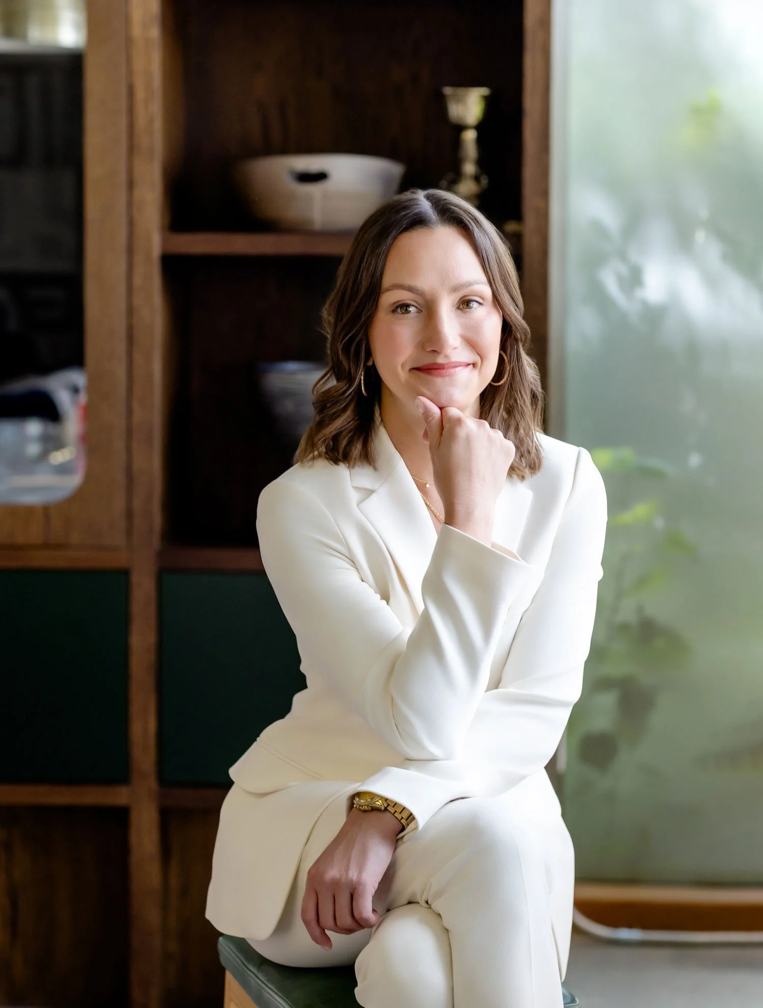 A woman in a white suit sitting on a chair, smiling at the camera, with a wooden shelf and greenery in the background.
