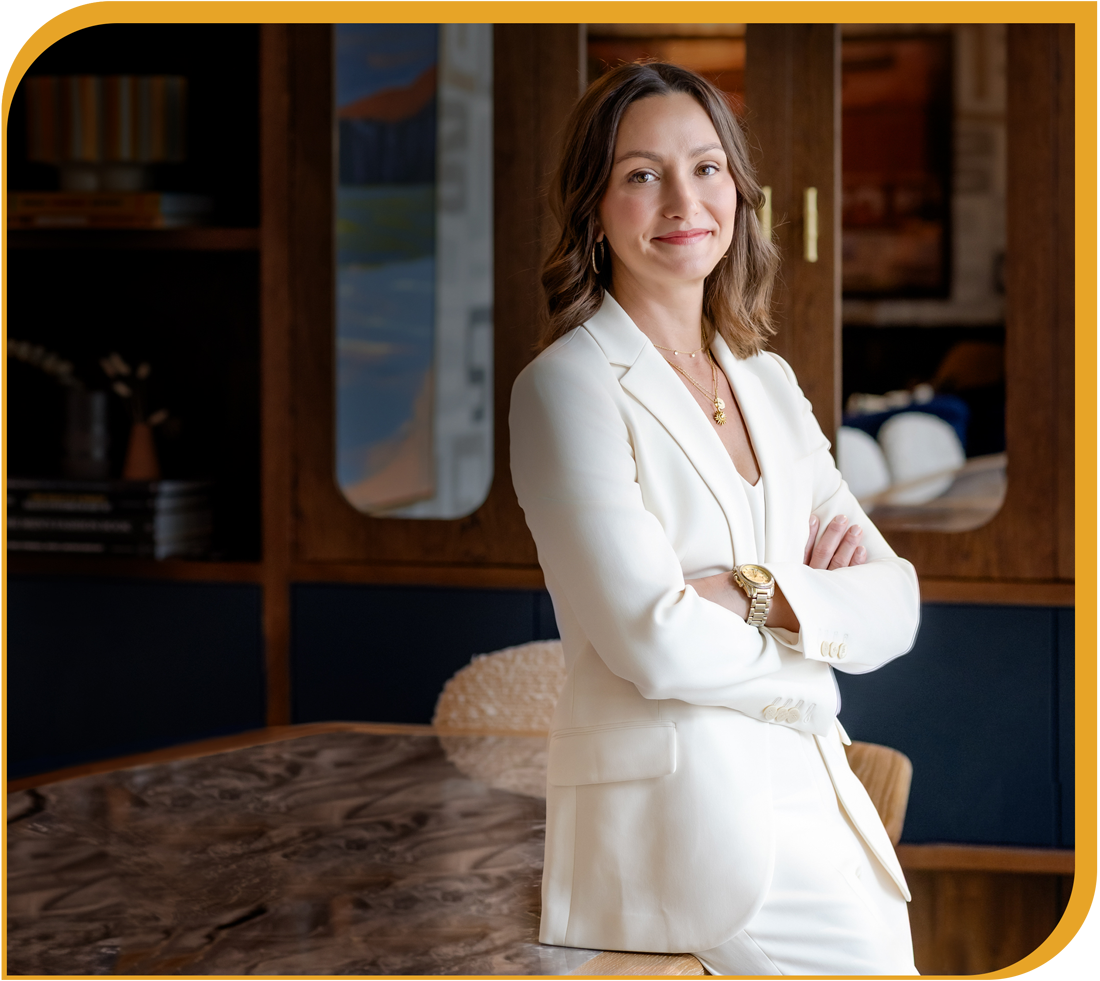 A woman with shoulder-length brown hair wearing a white suit and gold jewelry standing with arms crossed in a modern, stylish indoor setting.
