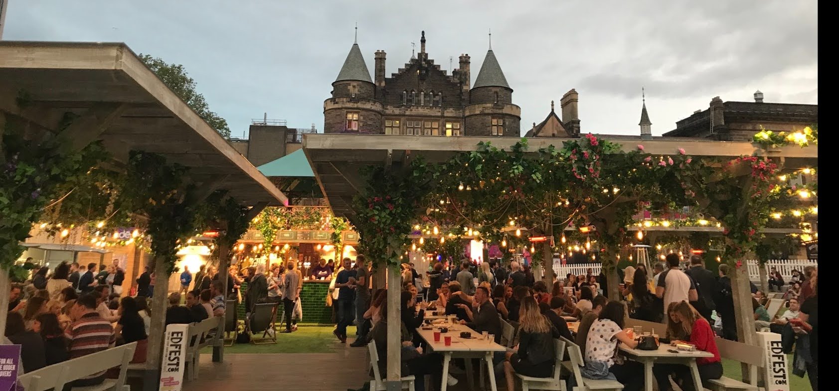 People sitting at white tables under vine-covered awnings with a gothic building in the background