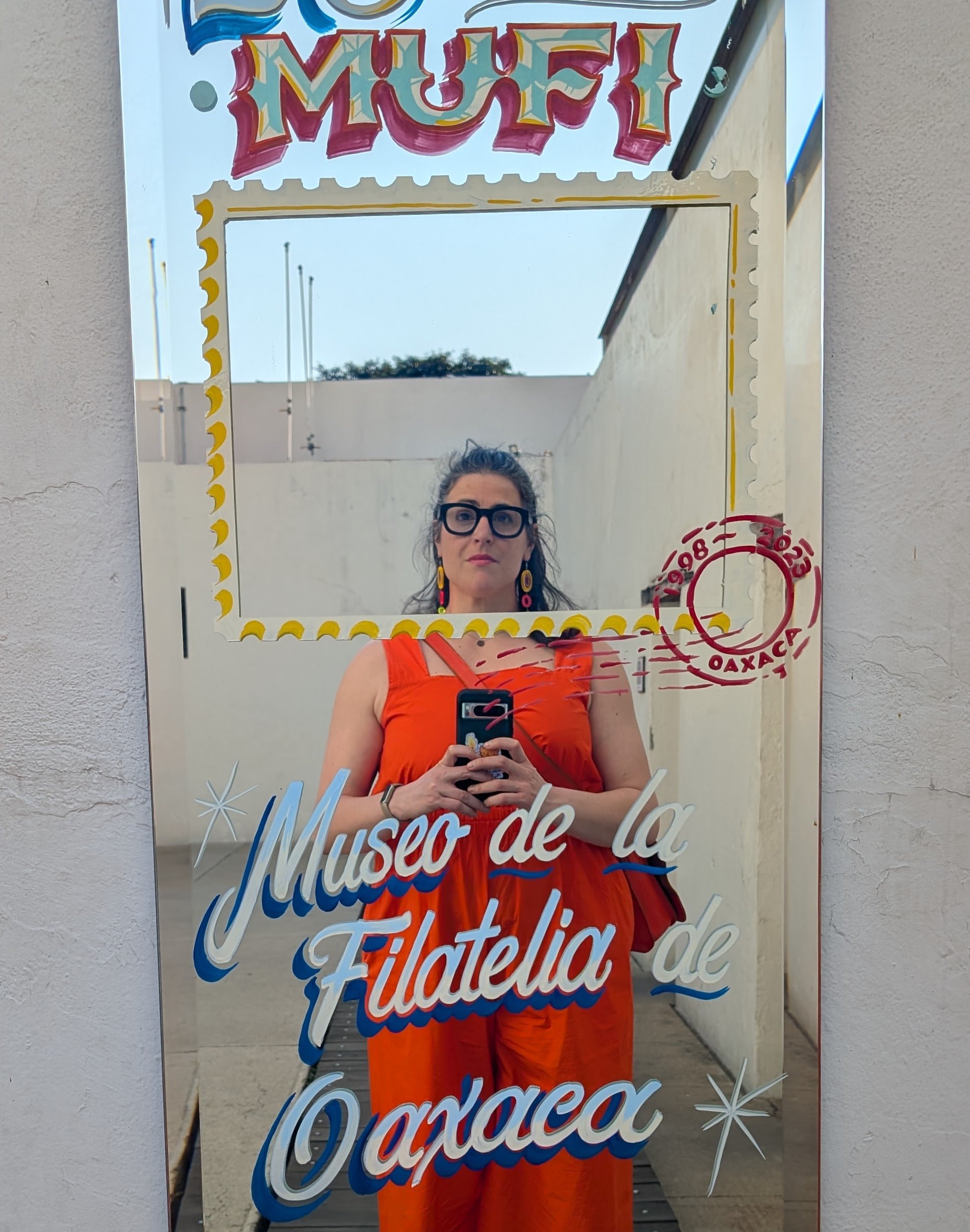 A mirror with colorful writing and decorations, reflecting a woman dressed in an orange top and skirt, with glasses and earrings, standing outdoors against a wall with a cloudy sky visible above.