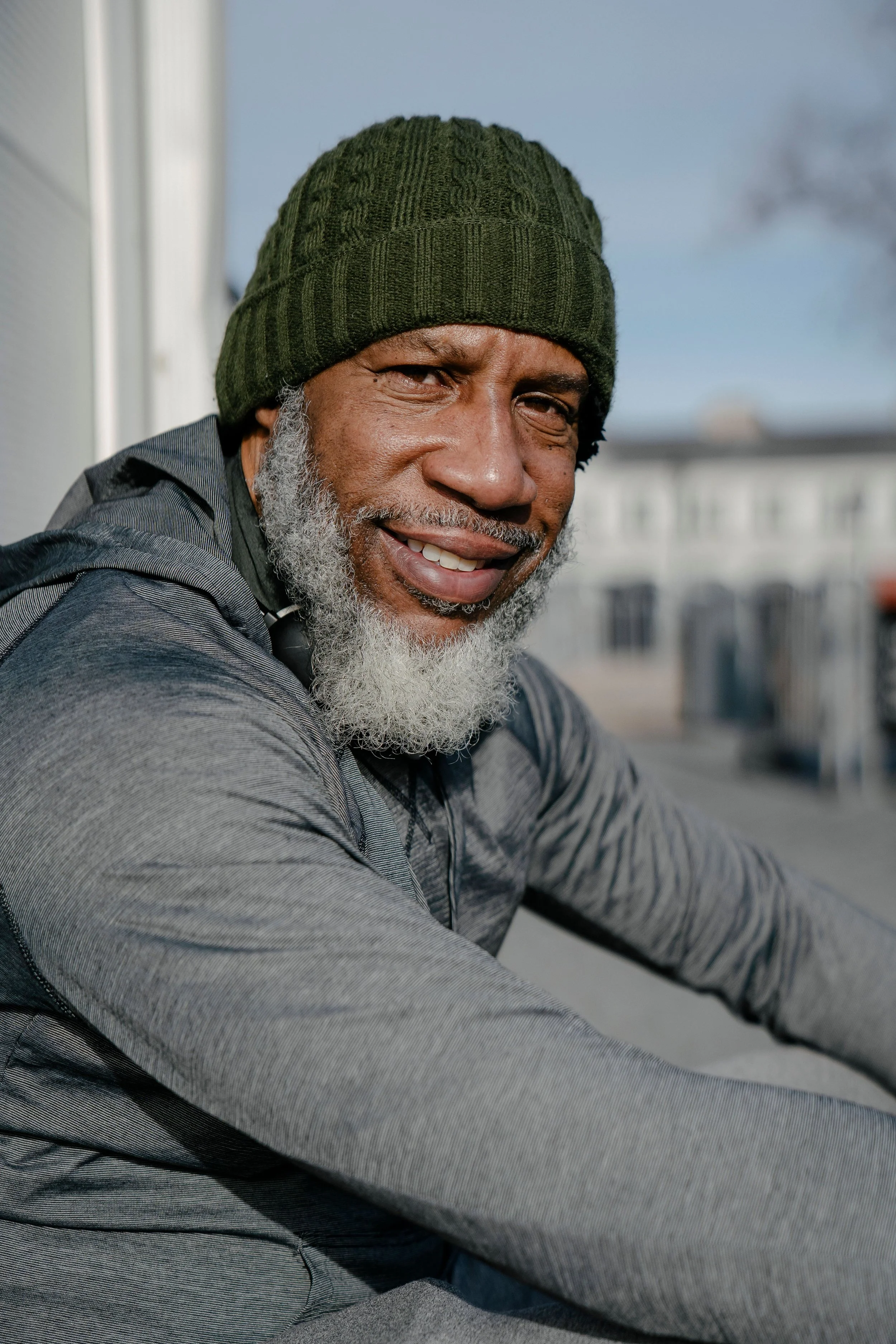 Close-up of a smiling middle-aged African American man wearing a green knit beanie and gray striped long-sleeve shirt, sitting outdoors.