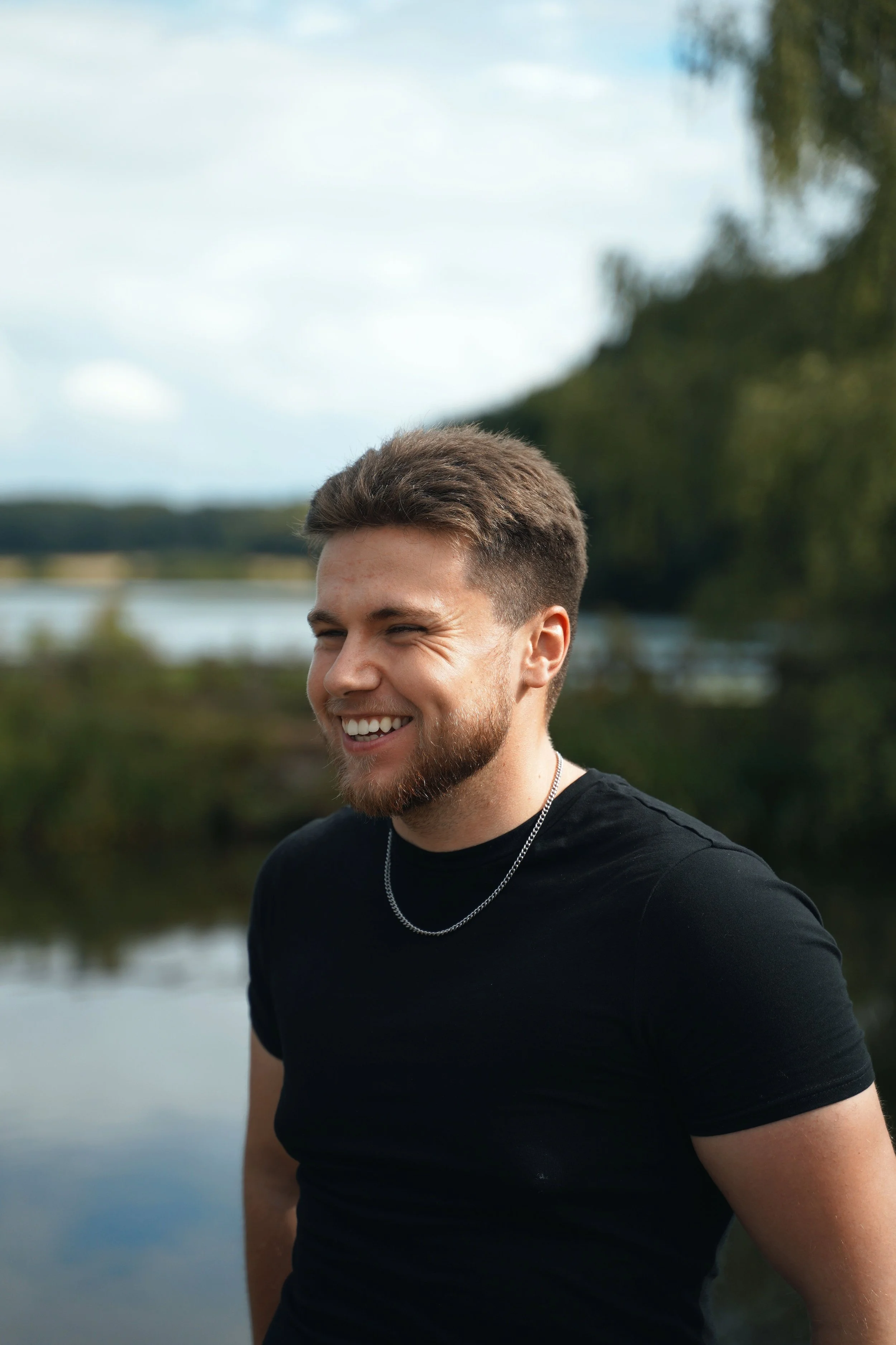 A smiling young man with a beard and short brown hair wearing a black t-shirt and a necklace, standing outdoors near a body of water with trees and a cloudy sky in the background.