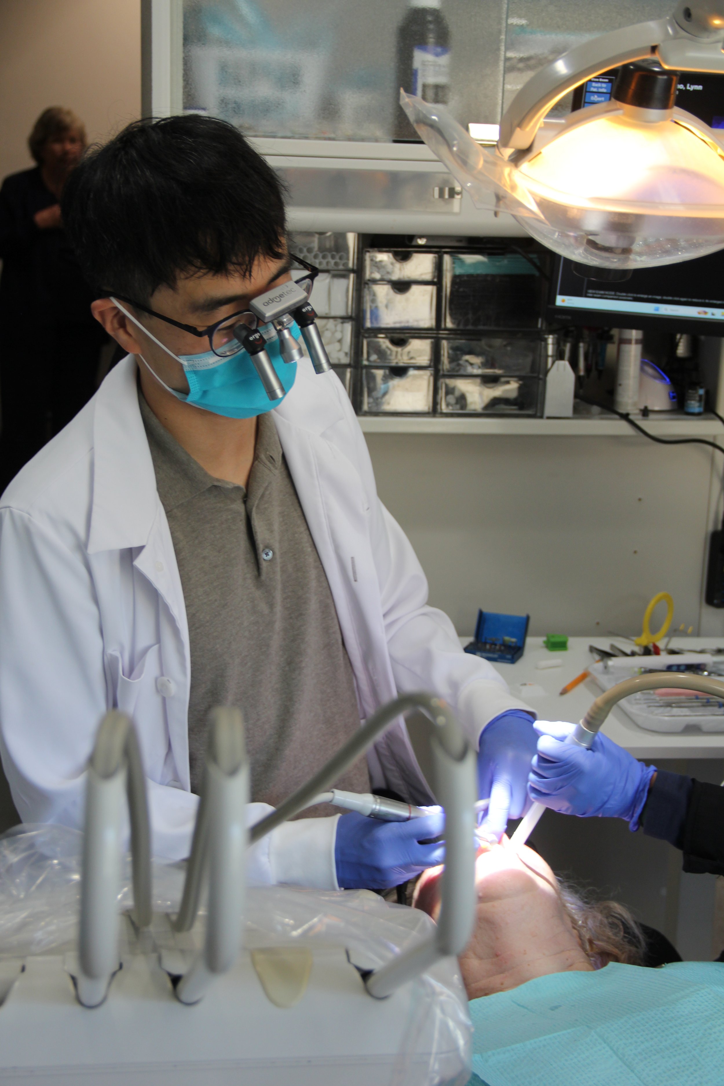 A medical professional using surgical tools on a patient in an operating room, wearing a mask and magnifying glasses.