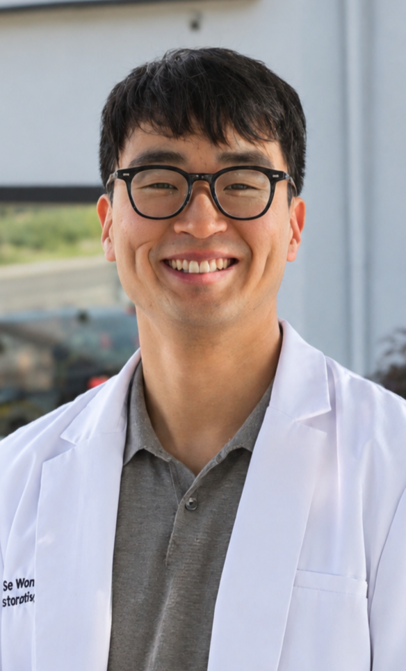 A smiling, confident male doctor wearing glasses and a white lab coat standing outside.