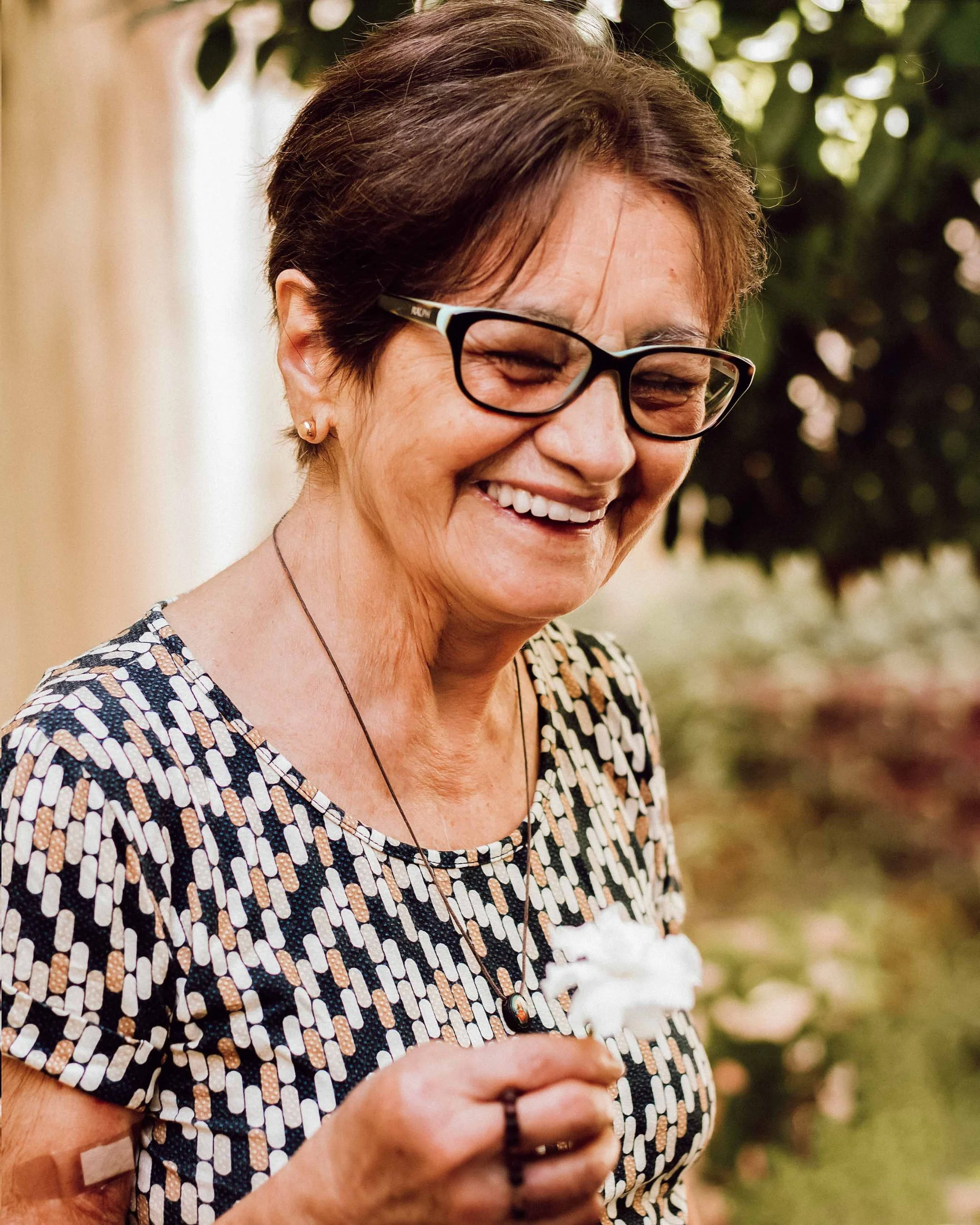 Close-up of a smiling woman with short brown hair, glasses, and a patterned dress, holding a white flower, outdoors with greenery in the background.