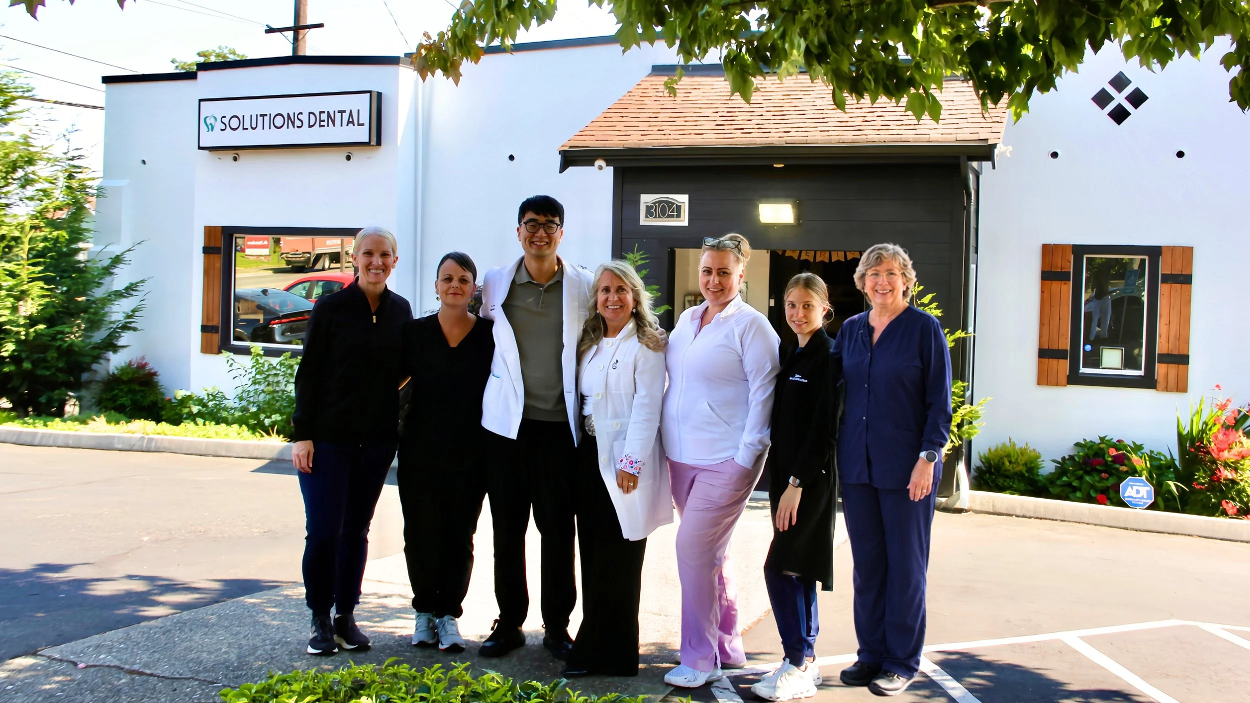 Group of seven medical professionals standing outside Solutions Dental clinic, smiling, in front of the building with a sign, surrounded by greenery.