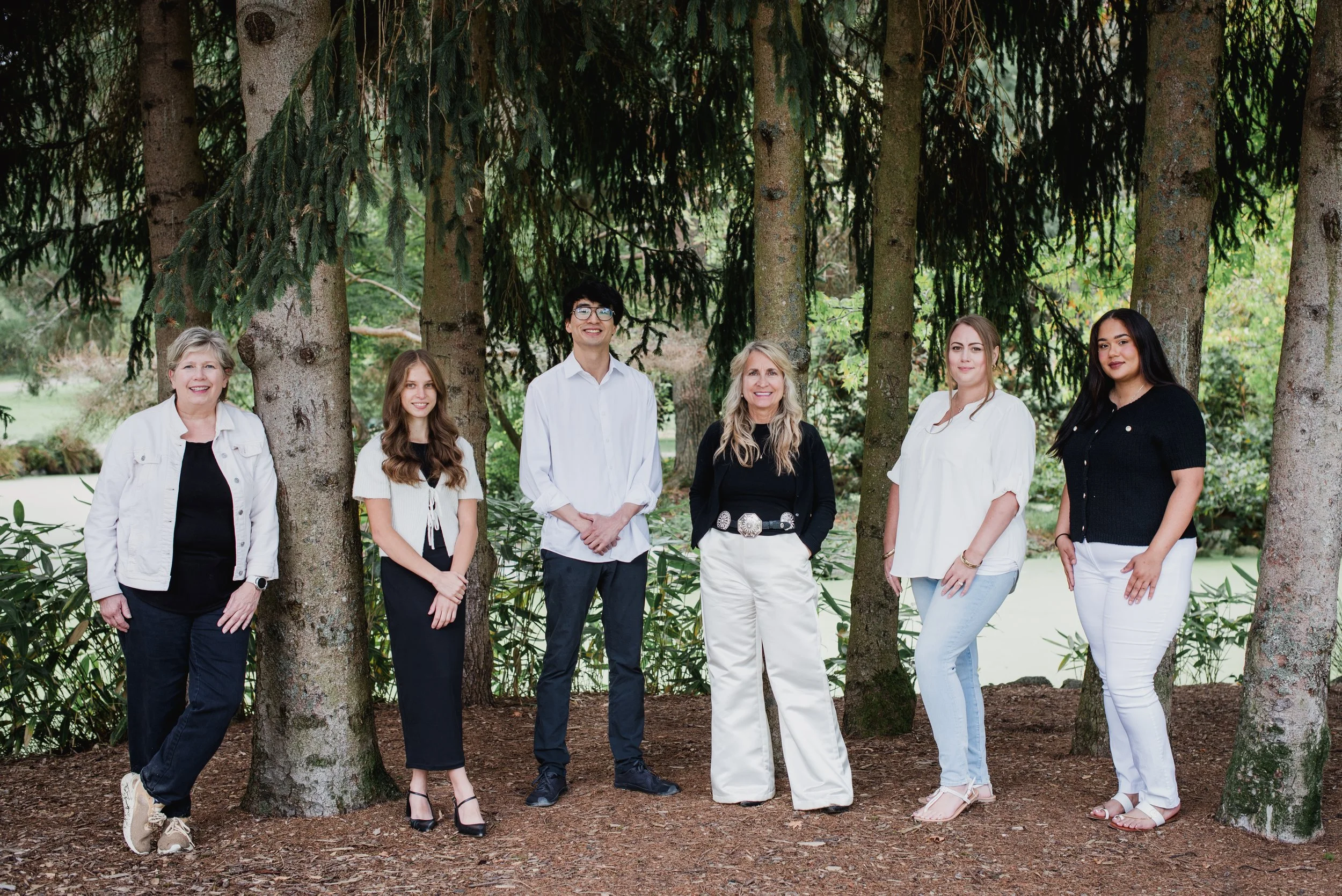 Group of seven diverse people standing outdoors among trees and greenery, smiling for the camera.