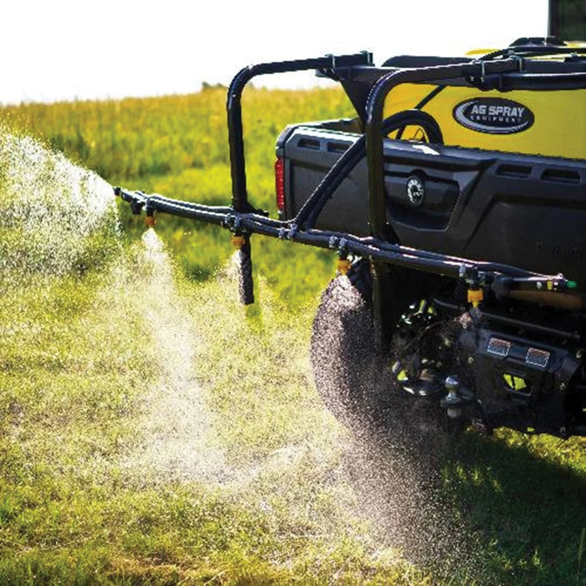 A yellow and black spray truck spraying pesticides or fertilizers on a green field in bright sunlight.
