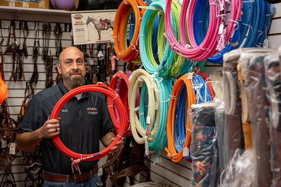 A man with a beard and dark shirt standing in a store aisle, holding a red tube. Behind him are colorful hoses hanging on a wall, with other horse tack and riding gear visible around.