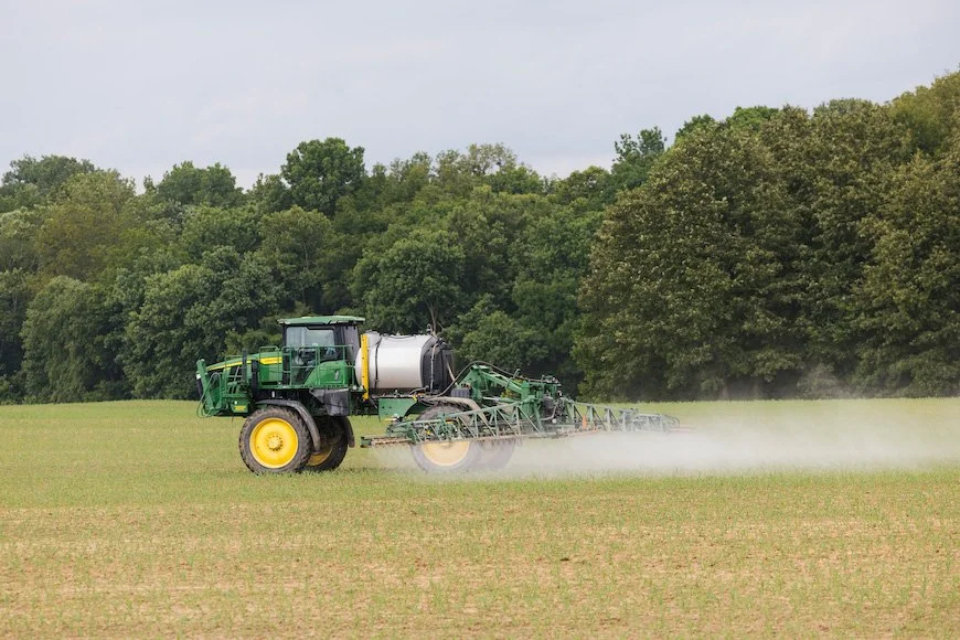 Green and yellow tractor spraying pesticide or fertilizer on a field with a backdrop of trees and a cloudy sky.