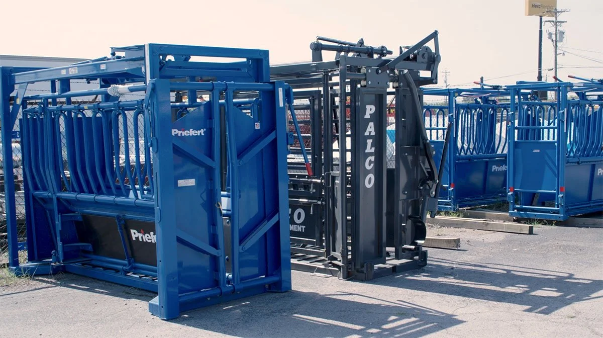 Metal cattle panels and gates stacked outdoors in a chain-link fenced lot, with some electric utility poles in the background.