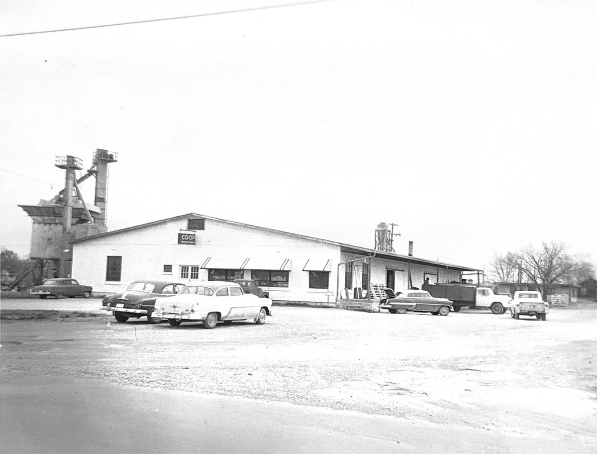 Black and white photo of a large warehouse with the 'CO-OP' sign, surrounded by several vintage cars parked in front.