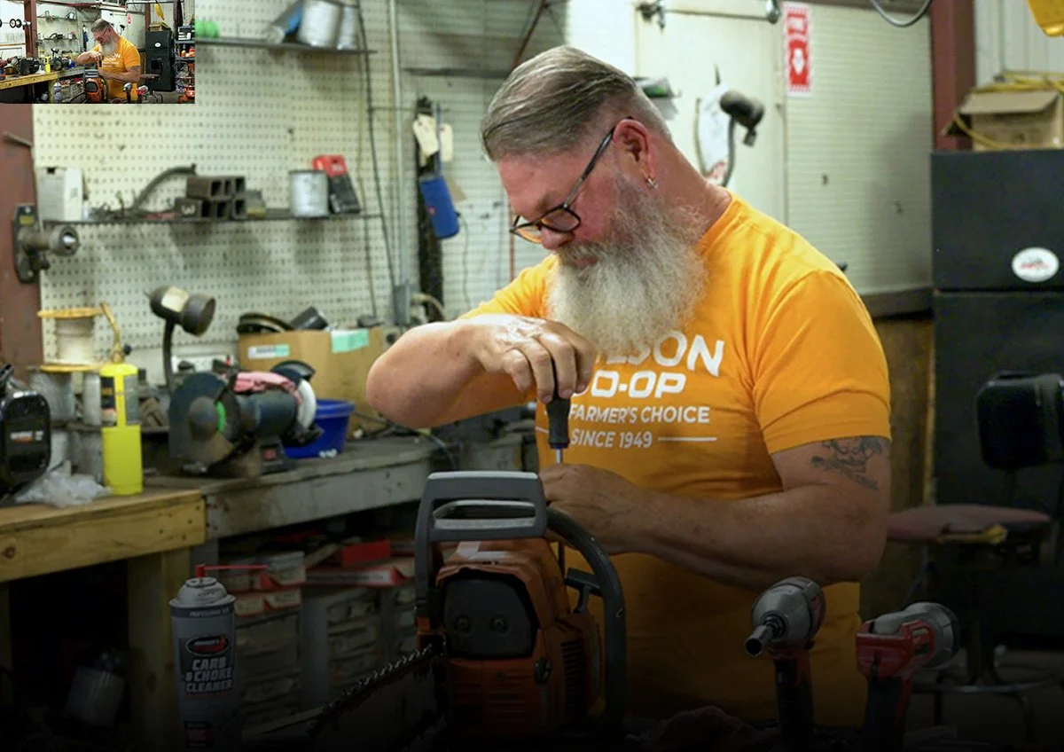 A man with a long beard, glasses, and earrings working on a chainsaw in a workshop filled with tools and equipment.