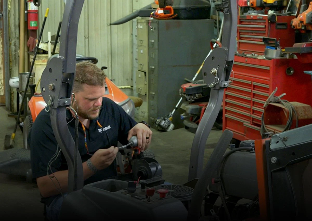 A man working on a lawnmower in a cluttered workshop with tools and equipment, including a red tool chest and a fire extinguisher in the background.