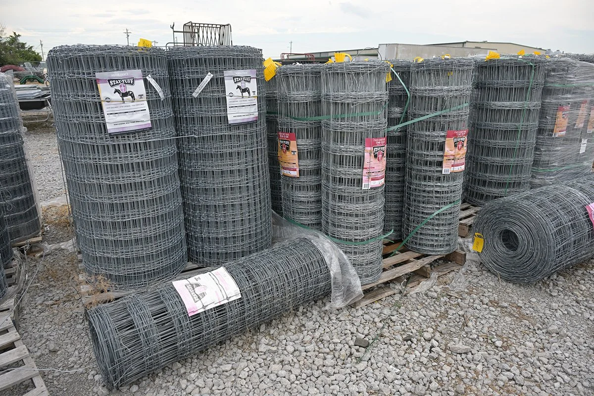 Stacks of rolled wire fencing panels and rolls on a gravel surface outdoors at a hardware or farm store.