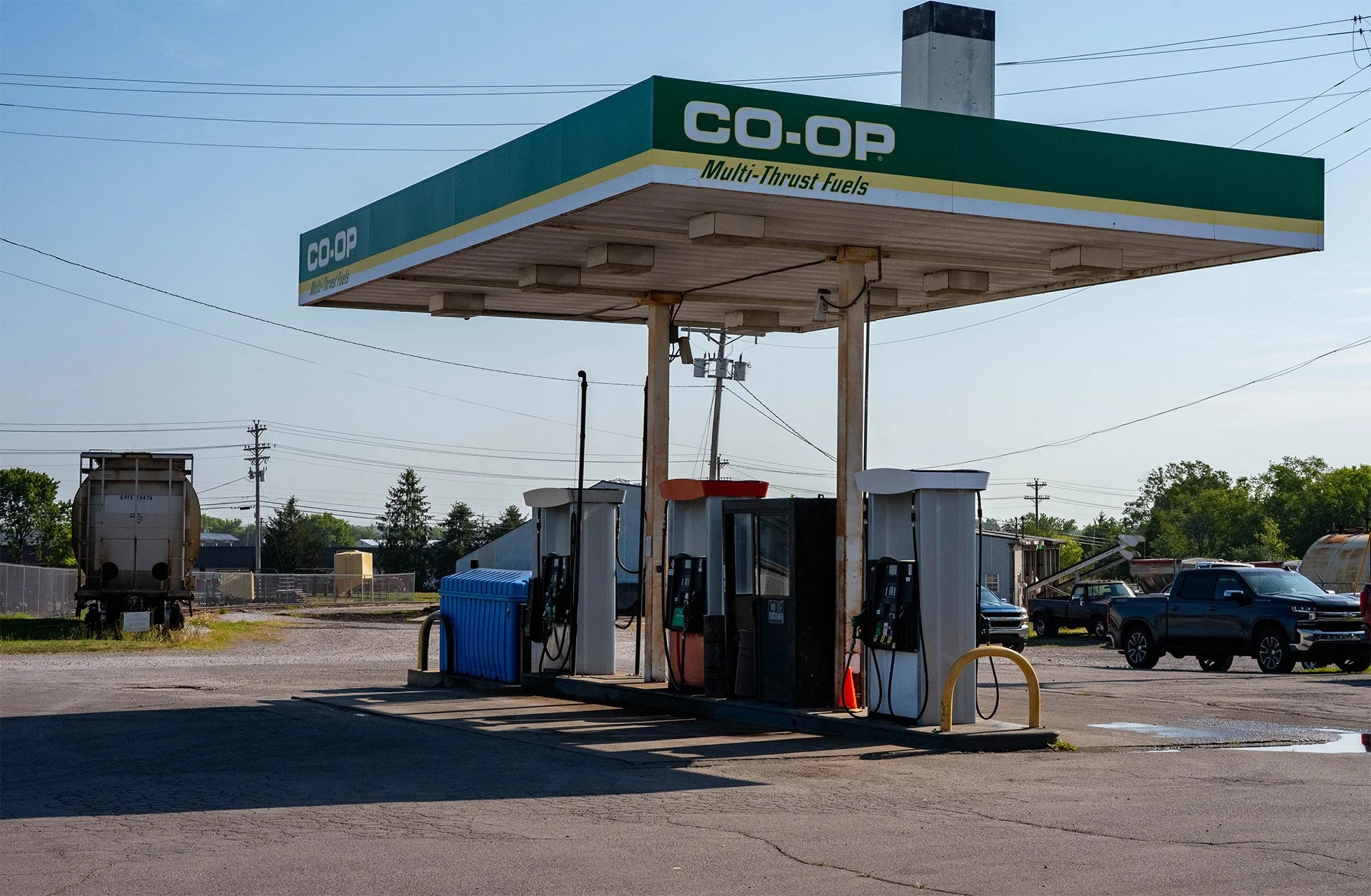 A gas station with a green and yellow canopy that reads 'CO-OP Multi-Thrust Fuels'. There are fuel pumps under the canopy, and a blue recycling bin is placed near them. In the background, there are vehicles, trees, power lines, and a large tank on the left side.
