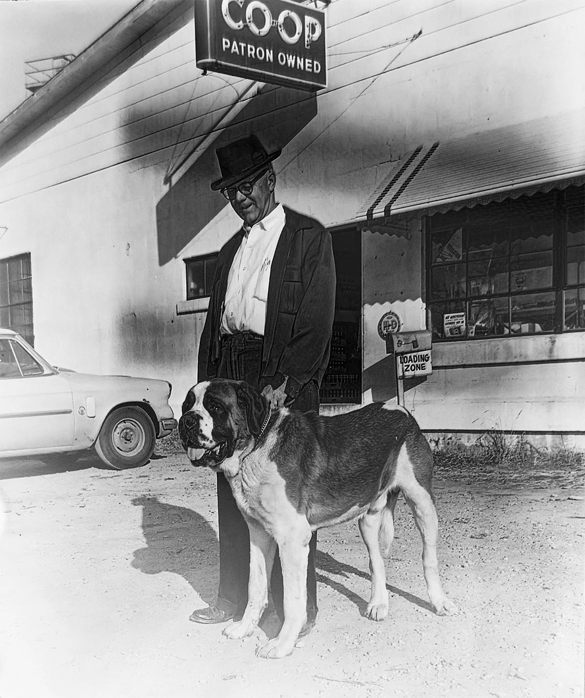 A man in a hat and glasses standing outside a store with a large dog, possibly a Saint Bernard, in front of him. The store has a sign that reads 'C.O.P Patron Owned,' and there is a car parked nearby. The photograph appears to be from the mid-20th century.