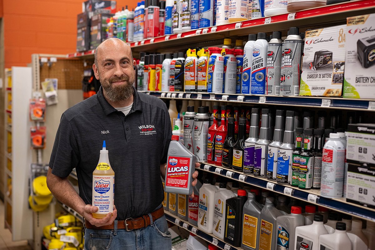 A man with a bald head and beard, wearing a black polo shirt with a company logo, standing in an automotive store aisle, holding bottles of Lucas oil products, including fuel injector cleaner and additive, with shelves stocked with various automotive fluids behind him.