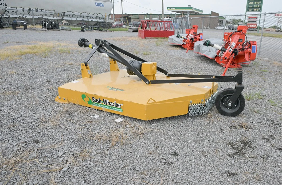 A Bush-Whacker turf mower for cutting grass, with a yellow deck and black handles, situated on a gravel lot with equipment and a chain-link fence in the background.