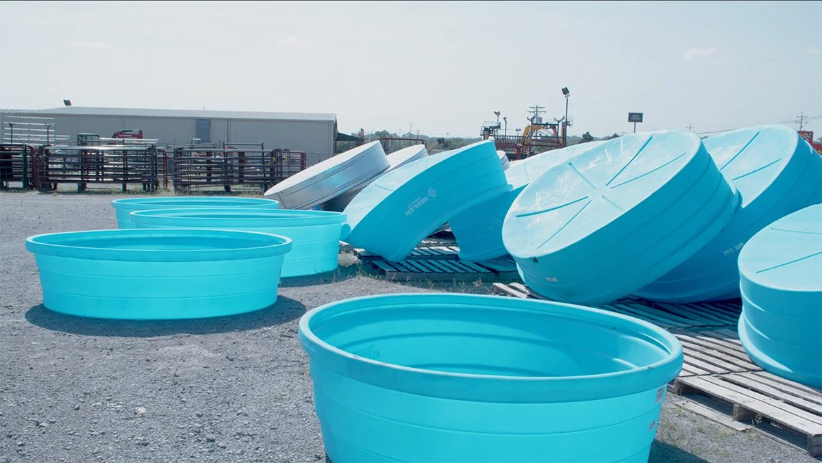Blue plastic kiddie pools and large round plastic tank lids stacked outdoors on gravel with a metal building and equipment in the background.
